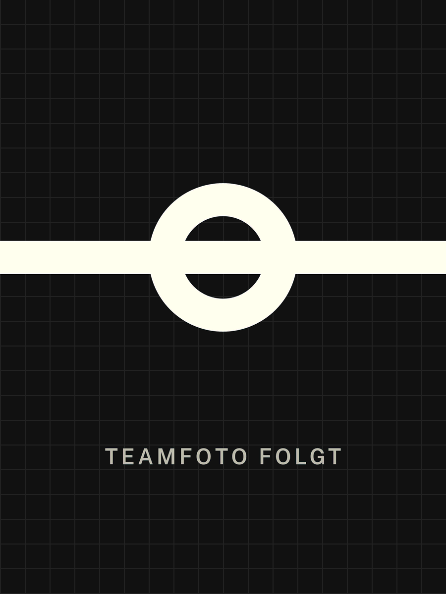 Gestalteter Platzhalter im Corporate Design: Ring mit durchgehender Achse auf graphitfarbenem Grund, darunter die Zeile „Teamfoto folgt“